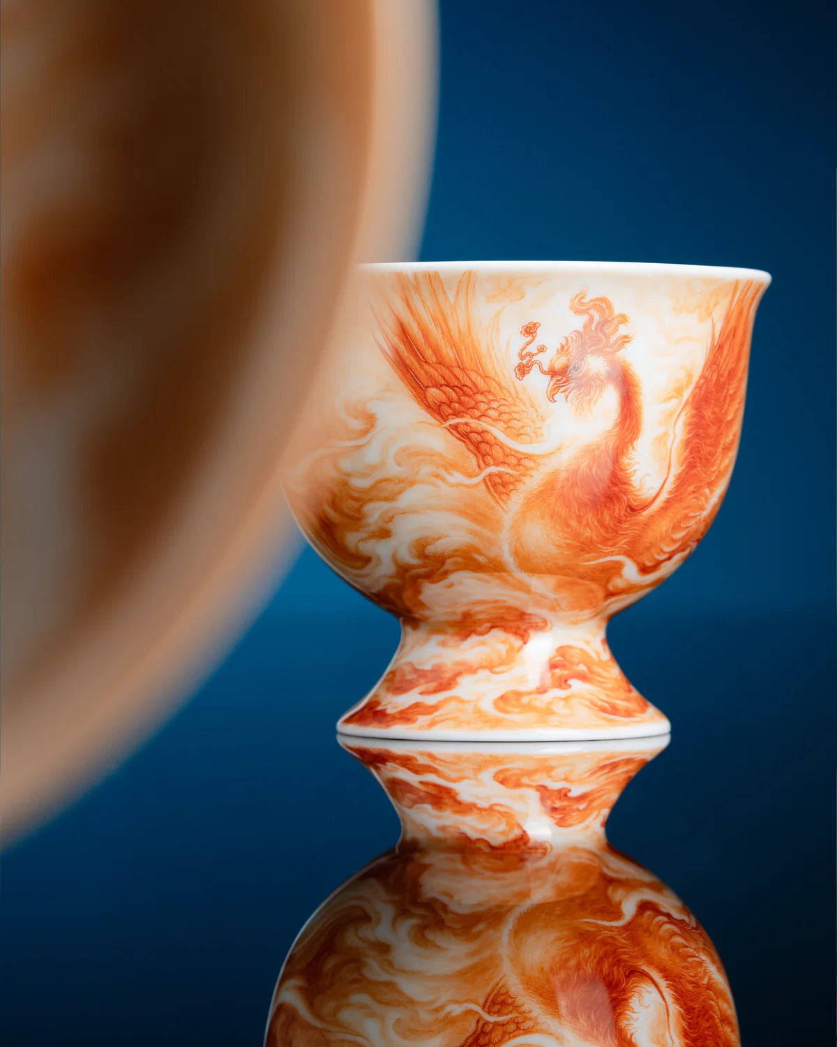 Orange-red phoenix motif on a porcelain cup reflected on a dark blue surface